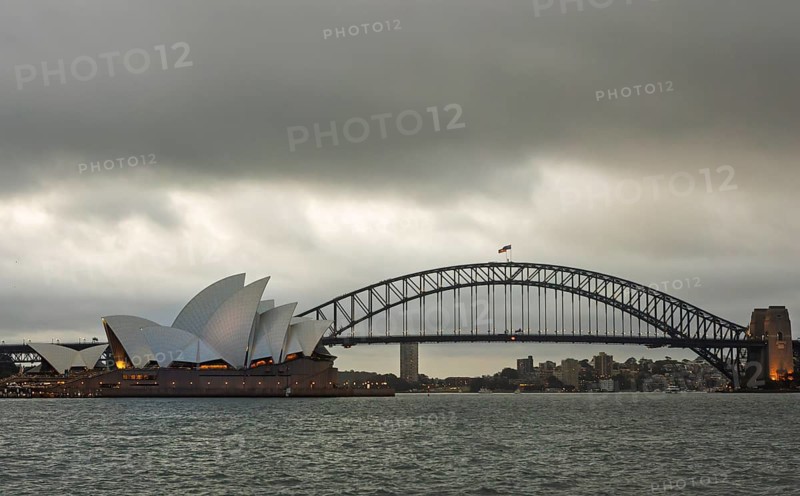 Moody sky over Sydney with the famed Opera House and Harbour Bridge in view. The still water reflects the iconic skyline, capturing a moment of calm in the bustling city