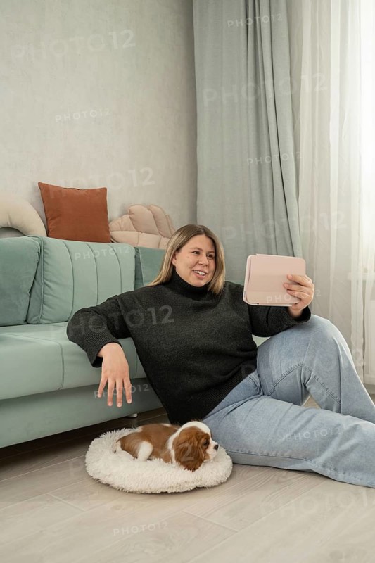 Woman relaxing at home with her adorable puppy resting nearby. She enjoys digital content on her tablet while sitting on the floor, showcasing a cozy living space