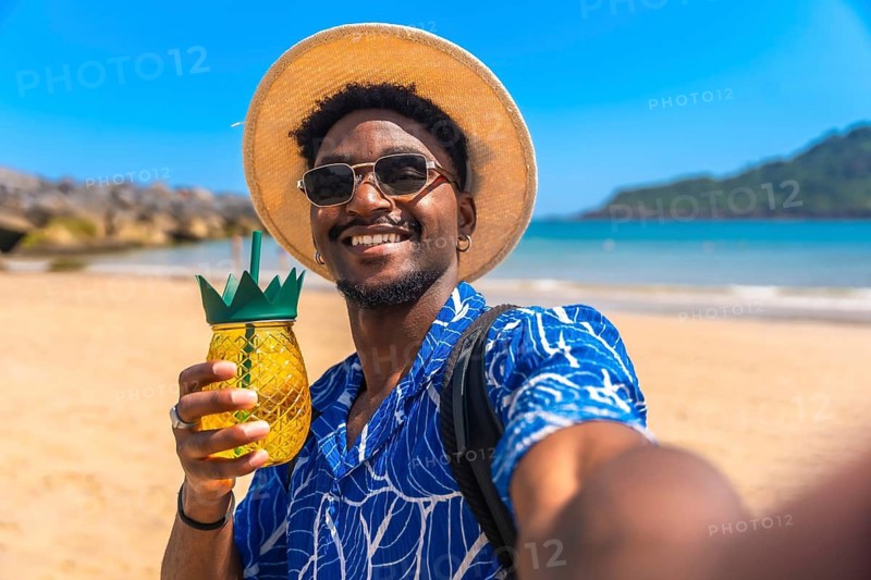 Young black man taking a selfie on a tropical beach while holding a refreshing pineapple cocktail