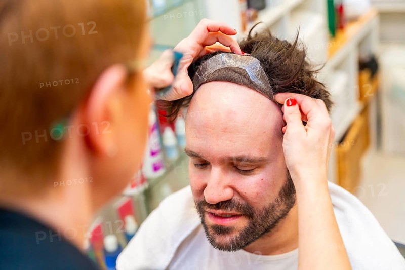 Close-up top view of a hairdresser attaching a capillary prosthesis carefully to a man's head