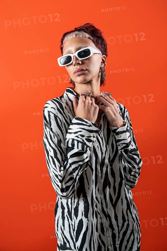 A fashion model poses confidently in a zebra-patterned blouse, adorned with modern accessories, against a bold orange backdrop in a studio with flash photography