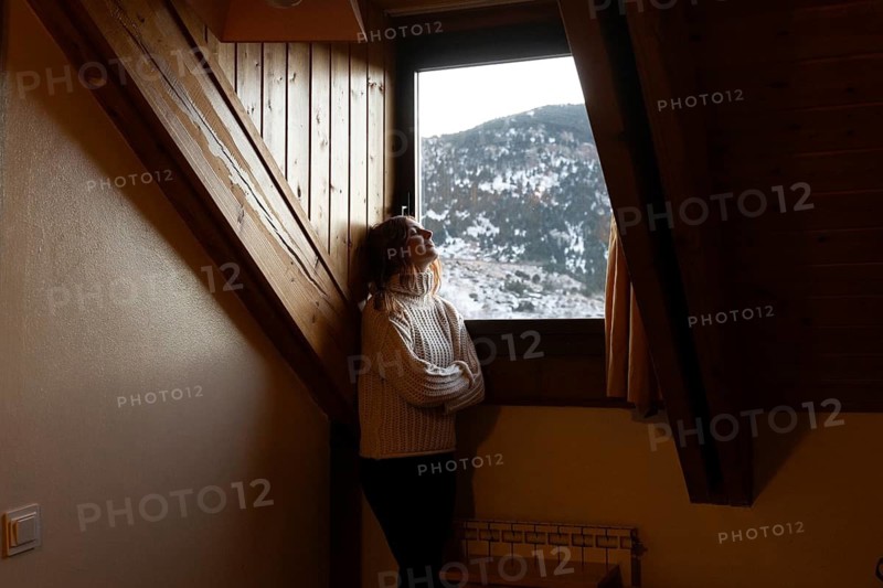 A woman in a cozy sweater stands by an attic window, gazing out at a snowy mountain landscape, embodying a peaceful winter moment indoors
