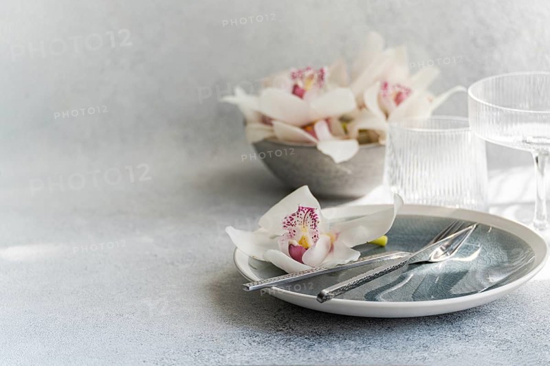 A serene table setting featuring delicate orchids on a pastel plate, accompanied by silver cutlery and textured glassware, conveying a modern and elegant dining ambiance