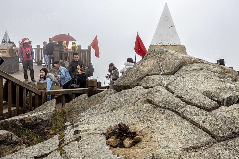 People on a viewing platform with triangular stone markings on rocks in foggy surroundings, The summit of Fansipan near Sa Pa in Vietnam