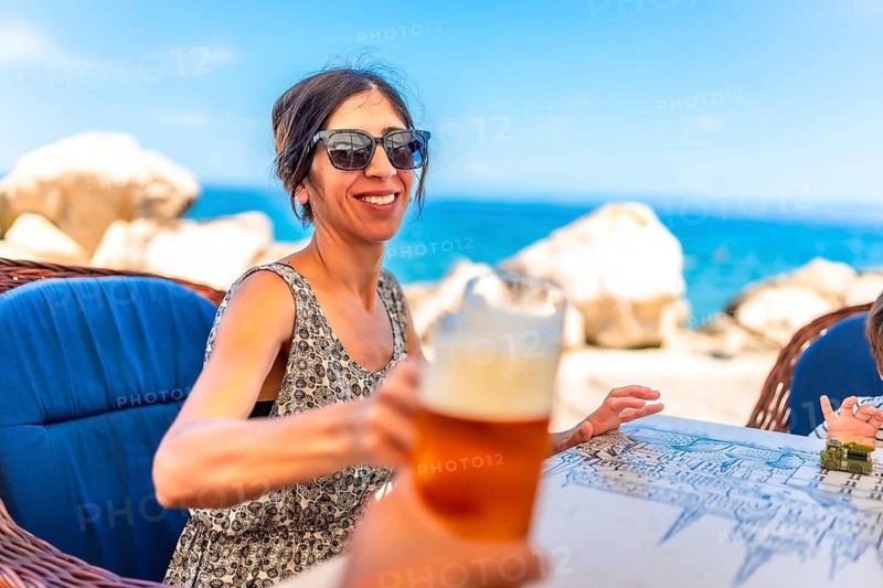 Woman toasting with a glass of beer at a seaside restaurant in piran, slovenia, enjoying a sunny day filled with laughter and relaxation