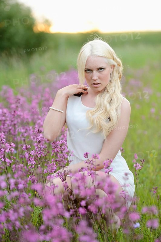 A blonde woman in a white dress sits among purple flowers in a natural field during the afternoon