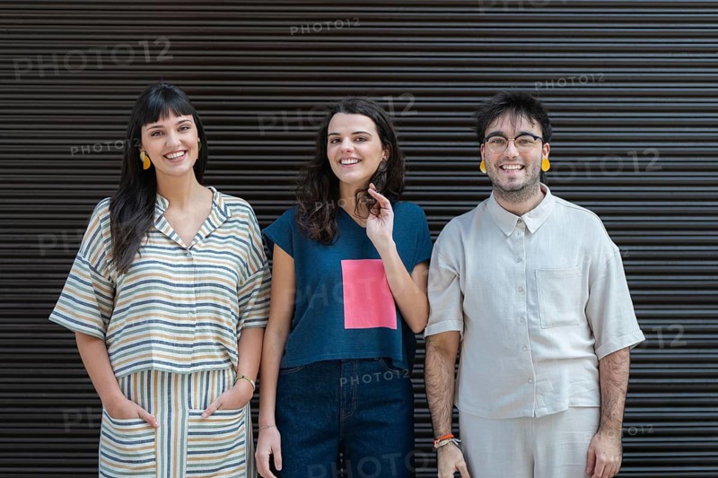 A diverse group of three friends stands together, smiling warmly at the camera. They each wear casual, trendy clothing while standing against a dark, striped backdrop