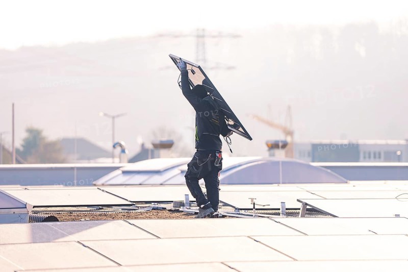 Person on roof lifts solar module surrounded by city and crane, clear daylight scene, PV installation at OBI Baumarkt, Mühlacker, Enzkreis, Germany