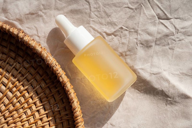 A cosmetic filled with yellow oil rests on textured fabric, next to a woven basket. Sunlit, it evokes simplicity, natural beauty, and tranquility