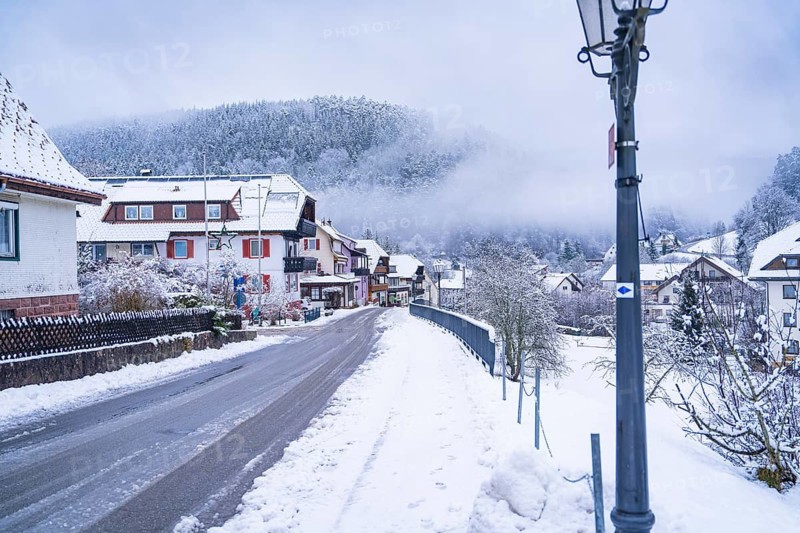Snow-covered village street with houses and lanterns, surrounded by fog-covered forests, Enzklösterle, district of Calw, Black Forest, Germany