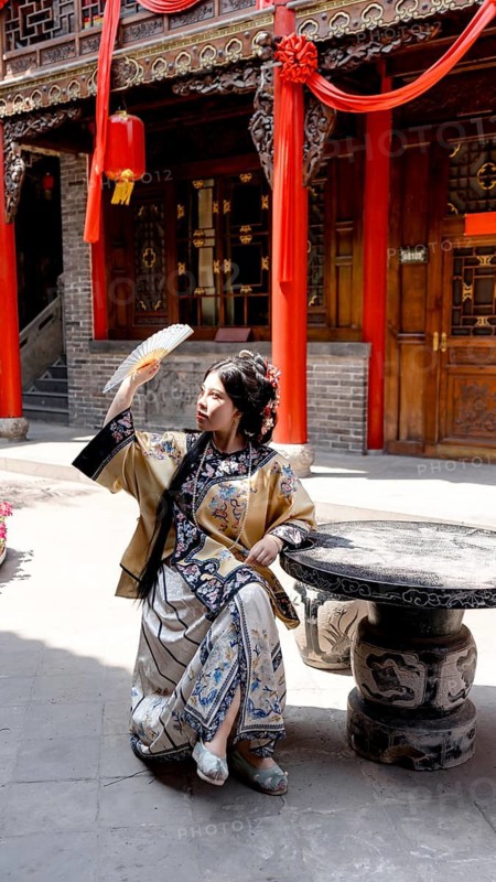 A woman dressed in elaborate Qing Dynasty attire sits gracefully outdoors in historic Pingyao, China, holding a fan under the bright sunlight, exuding elegance and tradition