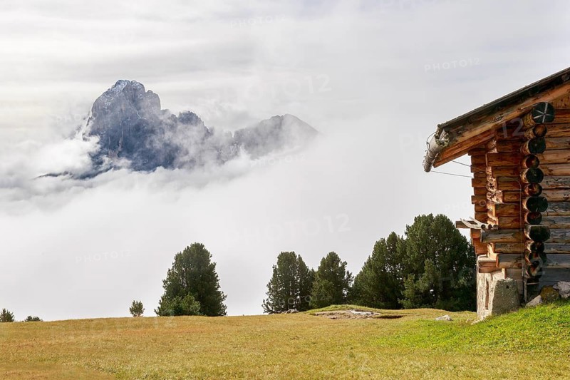 Rustic alpine hut on a green meadow, surrounded by mist and wild mountains in the background