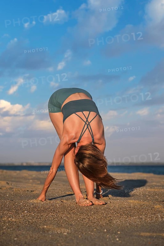 A woman practices Uttanasana, also known as Standing Forward Bend, on a sandy beach at sunset. The sun sets over the ocean