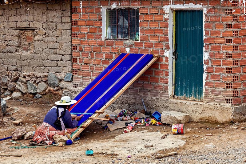 A woman in traditional clothes works on a blue loom in front of an outdoor brick wall, a woman weaves a cloth at Potosi on Cerro Rico in Bolivia