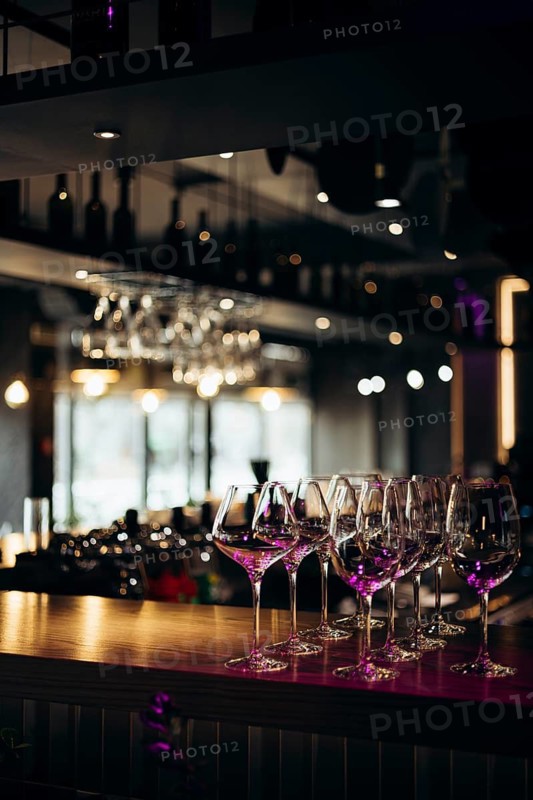 A collection of empty glasses elegantly lined up on a bar counter, illuminated by warm, ambient lighting. The scene sets a sophisticated and inviting atmosphere