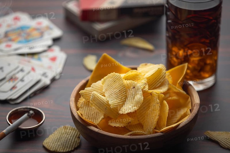 Bowl with chips, playing cards and cigarette next to drink on a table