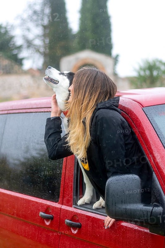 A woman leans out of a red car window, sharing a joyful moment with her dog. Both appear happy and relaxed, highlighting the bond between humans and their pets