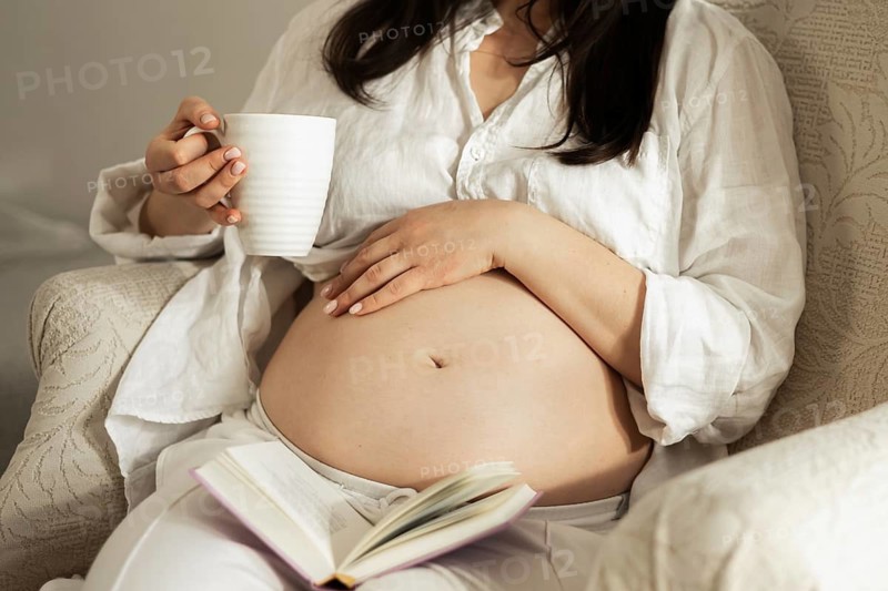 A pregnant woman enjoys a moment of relaxation, sipping tea and reading a book. Her hand gently rests on her belly, symbolizing anticipation and preparation for motherhood