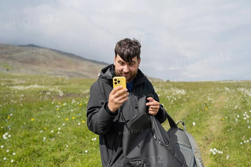 A lone hiker enjoys a picturesque mountain landscape, surrounded by vibrant wildflowers under a cloudy sky. Equipped for the journey, he captures his adventure on a smartphone