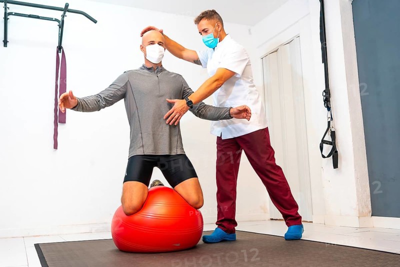 Client balancing on a red ball assisted by Physiotherapist with face mask. Physiotherapy with protective measures for the Coronavirus pandemic, COVID-19. Osteopathy, sports quiromassage