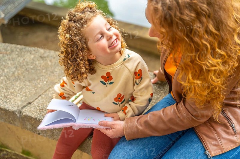 Adorable blonde girl smile while reading a book with her mother outdoors sitting in a park