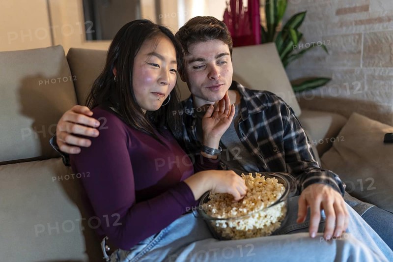 Asian woman and caucasian man enjoying a cozy evening at home, sharing a bowl of popcorn while sitting on a comfortable couch