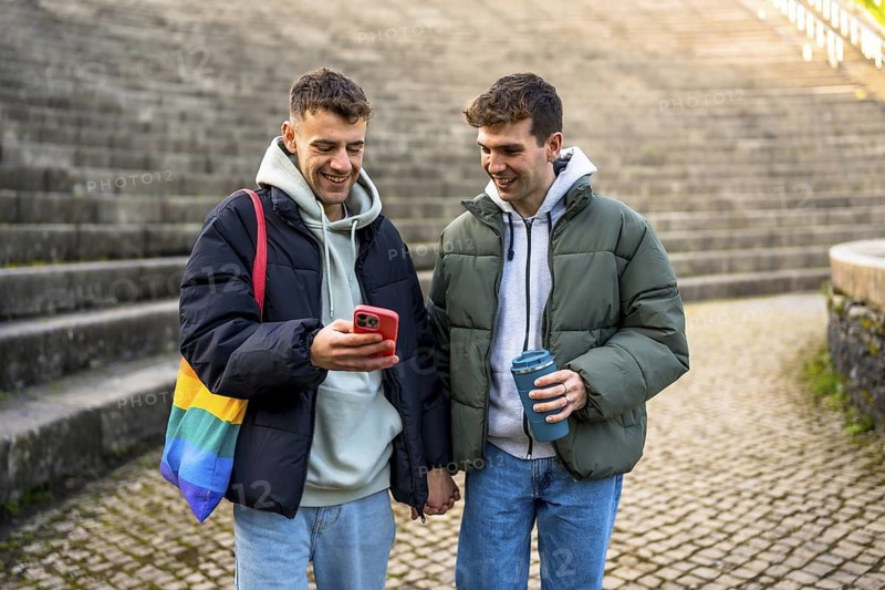 Happy gay couple walking hand in hand, smiling and checking their smartphone while enjoying a winter day outdoors in the city