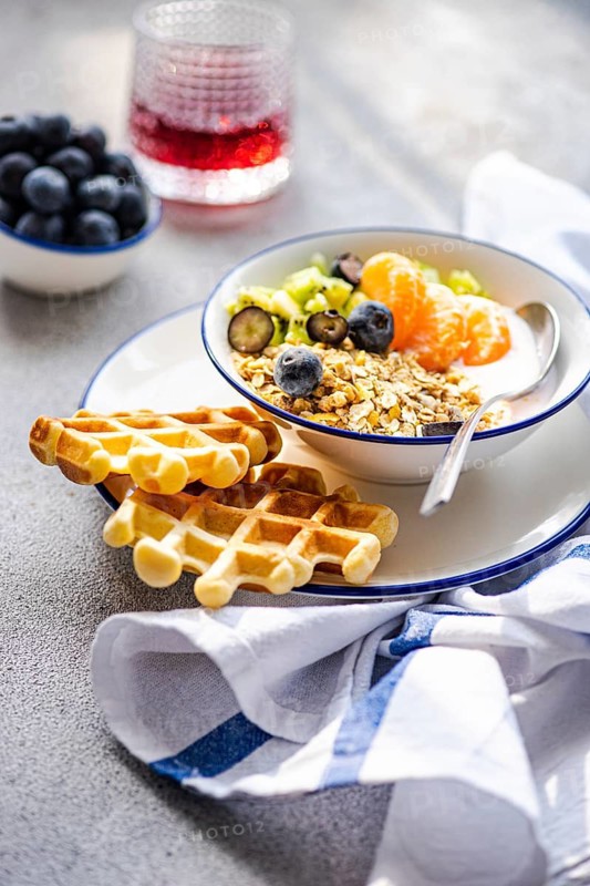 A vibrant breakfast scene featuring a bowl of granola with fresh fruits like blueberries and mandarins, served with waffles. A glass of juice and a bowl of blueberries are in the background