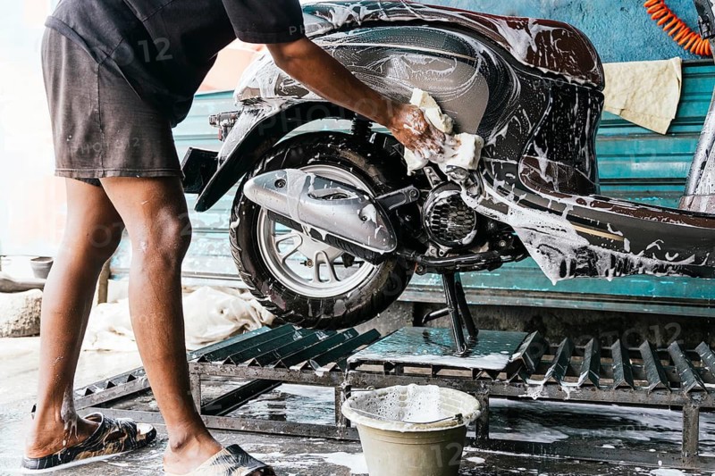 A man diligently cleans a scooter with soap and sponge, ensuring a thorough job. The scene captures attention to detail and care in motorcycle maintenance