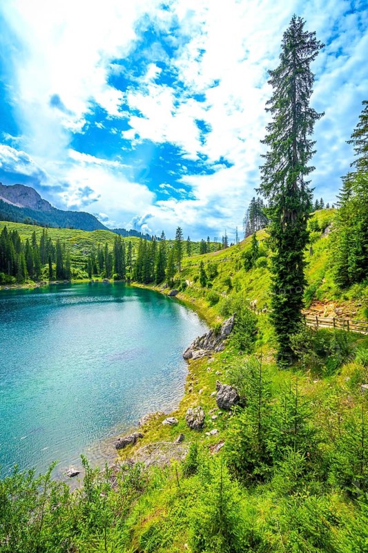 Alpine scenery reflecting on turquoise waters of karersee lake under a cloudy blue sky, surrounded by fir trees and green meadows