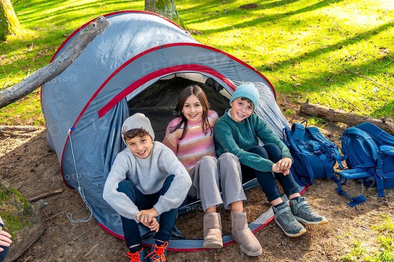 Three caucasian children sitting on a camping tent smiling at camera enjoying a day in the forest