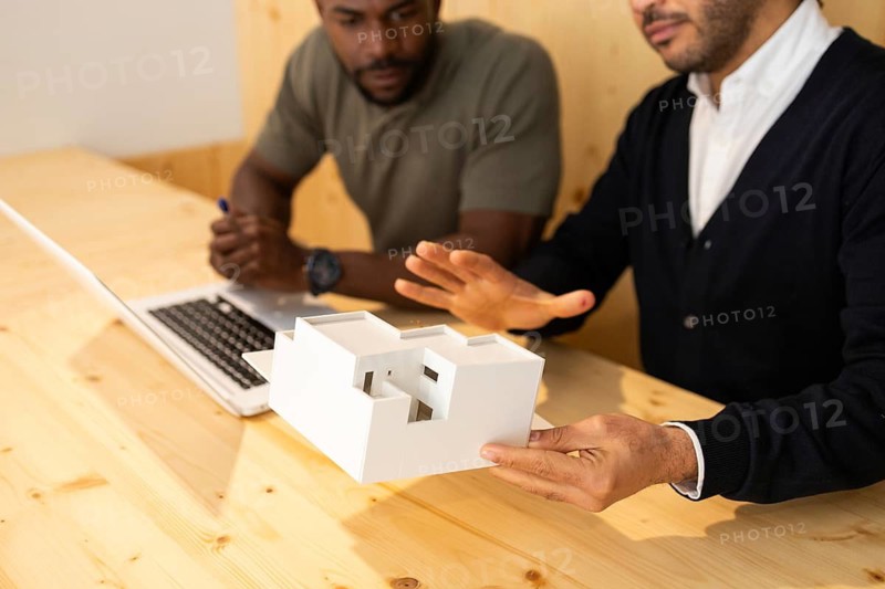 Two professionals in a modern coworking space in Barcelona discuss architectural concepts using a 3D model. Laptops and teamwork enhance their creative meeting