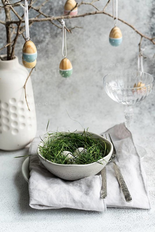 An elegantly set Easter table featuring a bowl of eggs nestled in fresh green grass, accompanied by a delicate arrangement of hanging artfully painted eggs and a sophisticated ceramic vase