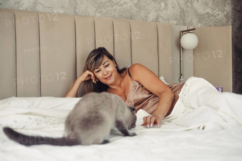 A woman in a satin nightgown lies comfortably on a plush bed, smiling as she plays with her pet cat. The cozy setting exudes warmth and relaxation in a serene bedroom