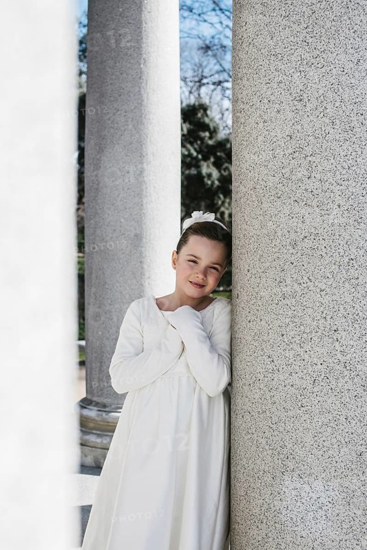 A young girl in a white First Communion dress stands between stone columns, smiling softly. The sunlight highlights her joyful expression on this special day