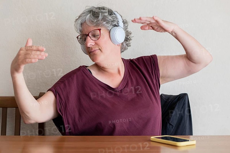 A senior woman wearing headphones dances joyfully at a table, embracing the music. A phone rests on the table. Her expression shows contentment and freedom