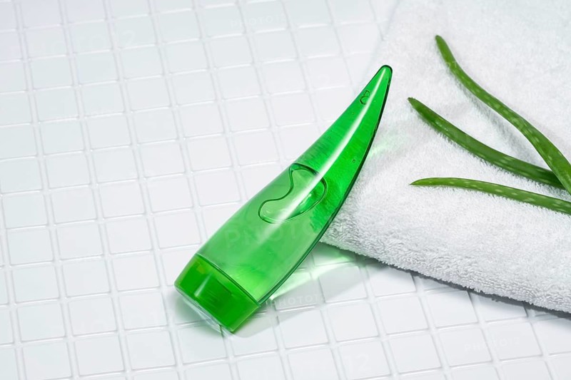 A green aloe vera gel tube rests next to aloe leaves on a white fluffy towel, all arranged on square bathroom tiles