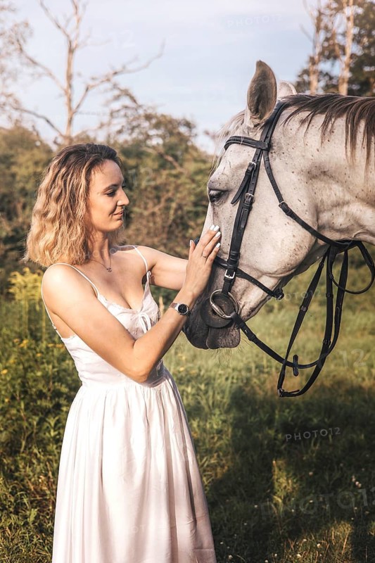 A young woman in a white dress gently touches a horse face, set against a lush green background. The scene captures a peaceful moment of connection and trust