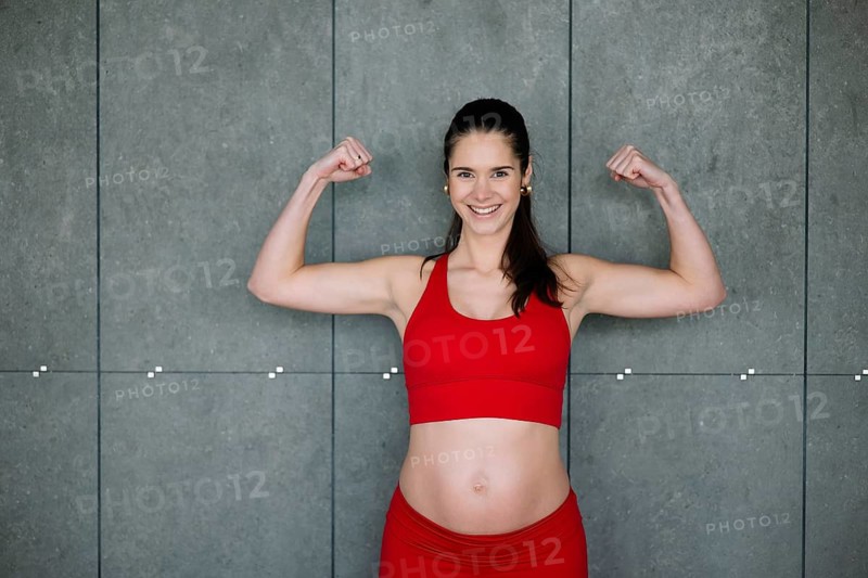 A cheerful pregnant woman confidently flexes her muscles while wearing a red yoga outfit, showcasing strength and positivity during pregnancy against a minimalist gray background