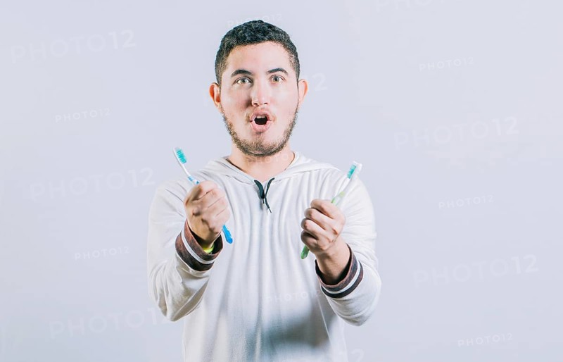 Amazed teen guy holding two toothbrush looking at camera isolated. Surprised people holding toothbrushes isolated