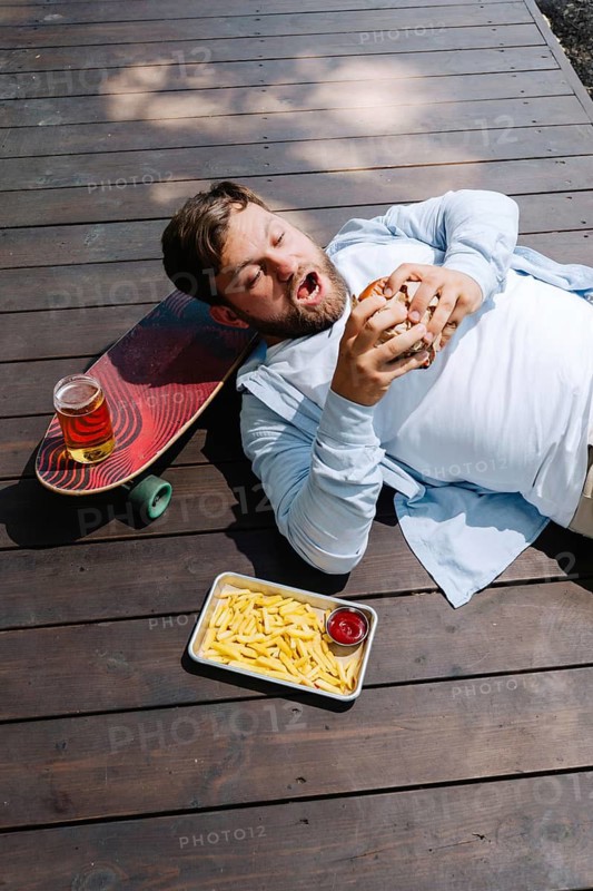 A man enjoys a burger, fries, and a drink while lounging outdoors on a wooden deck. A skateboard nearby adds a relaxed, adventurous vibe to the scene