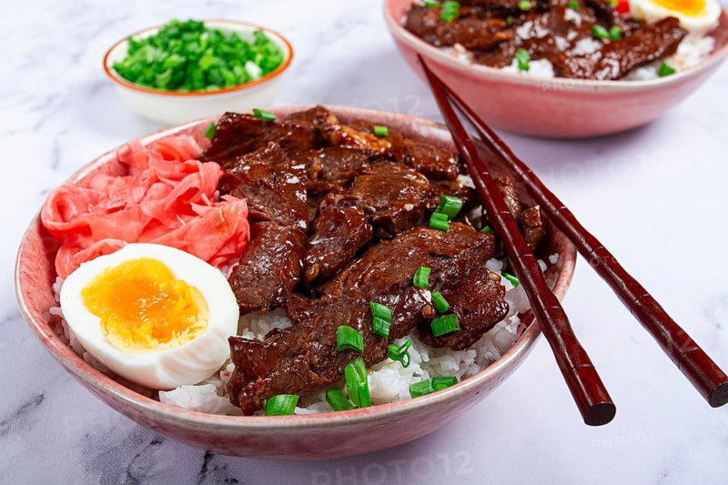 Gyudon Japanese beef with rice, pickled ginger, boiled egg slice, garnished with green onions, in a bowl with chopsticks