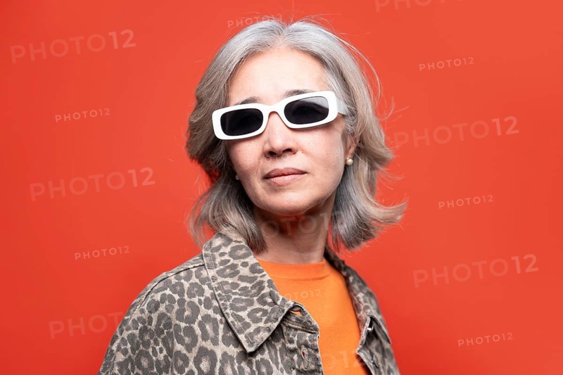 Asian senior woman with stylish sunglasses and a leopard print jacket, exuding confidence and personality against a vibrant orange background, embodying modern elegance