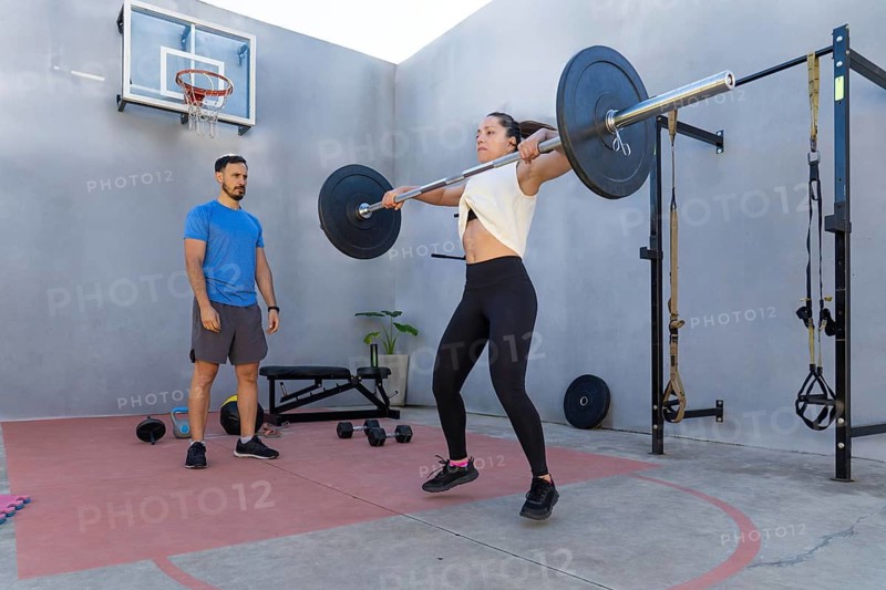 Young woman performing a power clean exercise with a barbell outdoors under the supervision of her personal trainer, demonstrating strength and determination in a functional training area