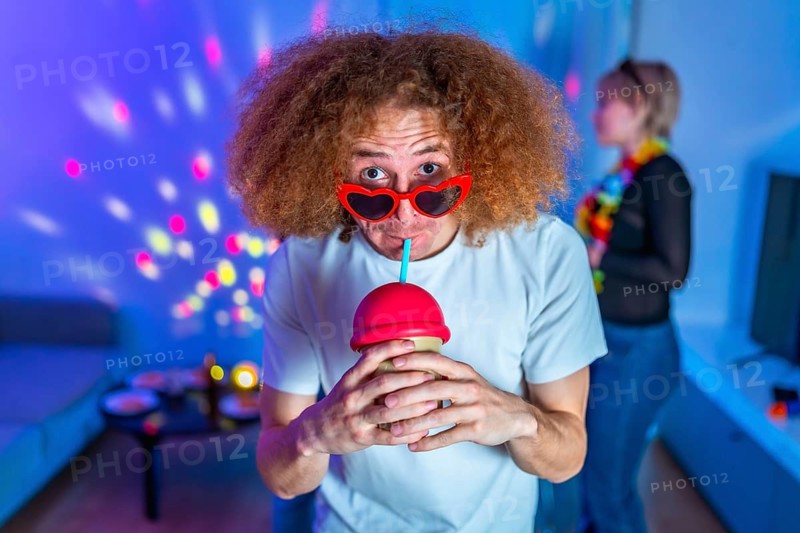 Ginger man wearing heart shaped sunglasses, enjoying a colorful cocktail while celebrating at a lively disco party with friends