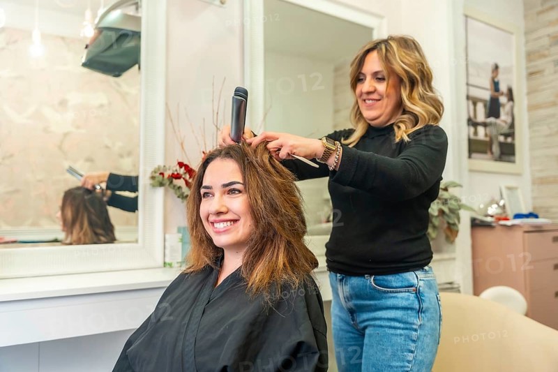 Hairdresser styling smiling client's long brown hair with a flat iron in a bright beauty salon, providing professional care and an enjoyable experience