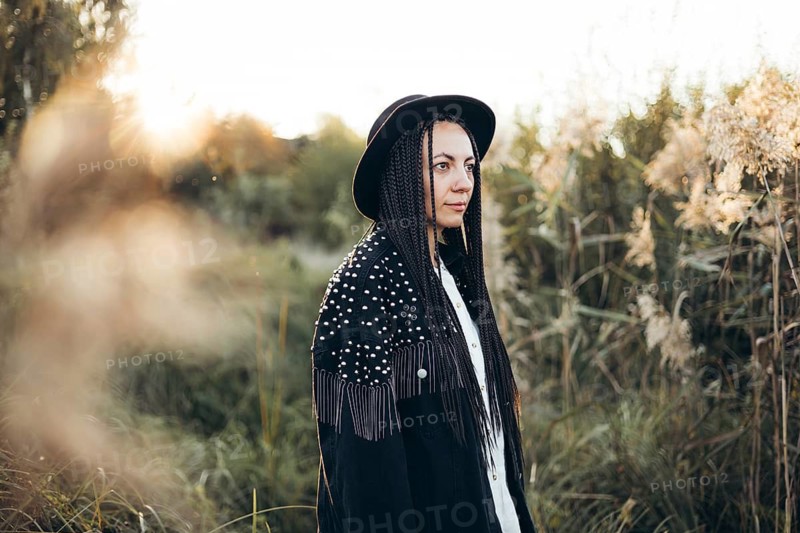 A woman stands in a serene field at sunset, wearing a stylish hat and braided hair. The warm glow of the sun creates a peaceful and contemplative atmosphere