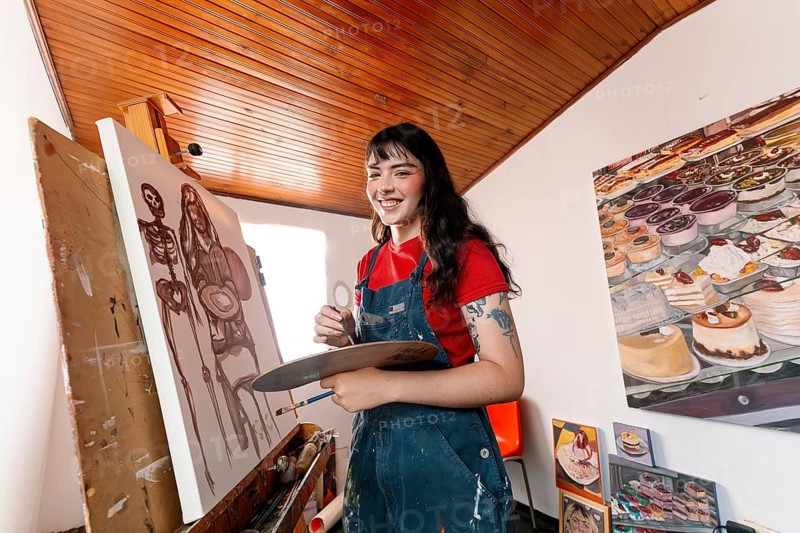 A young Latin woman joyfully paints on a canvas in her cozy home studio, surrounded by creative artworks. She holds a palette and brush, embodying artistic passion and creativity
