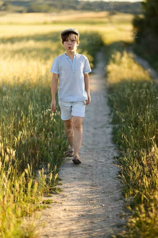 A young boy walks alone on a narrow dirt road bordered by wild grasses and fields during a tranquil summer evening. The warm sunlight highlights his thoughtful expression as he strolls in a casual outfit