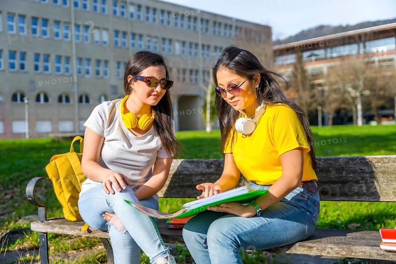 Multi-ethnic students with headphones looking at homework sitting on the bench of a campus